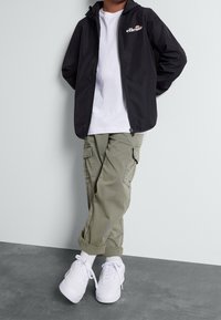 Zwarte waterdichte jas met capuchon, wit logo, wit T-shirt, olijfgroene cargobroek met zakken, en witte sneakers op grijze vloer.