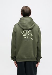 Πράσινη κουκούλα φούτερ από μαλακό ύφασμα, με μεγάλο λευκό γραφικό στην πλάτη και μικρό λογότυπο Nike πάνω από το στρίφωμα.