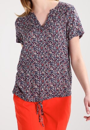 Mujer con una blusa de manga corta con estampado floral azul marino con un lazo delantero y pantalones rojos brillantes sobre un fondo liso.