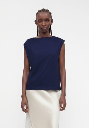 Femme aux cheveux tressés portant un haut bleu marine sans manches et une jupe en satin blanche avec des détails en dentelle, debout devant un fond uni.