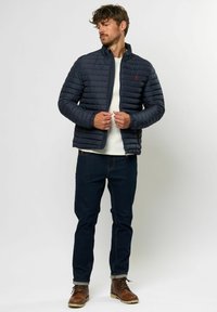 Marineblå puffjakke med horisontal quiltet mønster, høy krage og frontlukking med glidelås. Brukt med lys genser, mørke jeans og brune støvler.