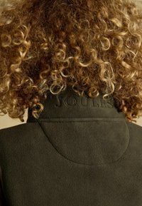 Zelená fleecová vesta s vysokým golierom, na zadnej strane s vytlačeným logom "JOULES". Tkanina má mäkký, textúrovaný vzhľad.