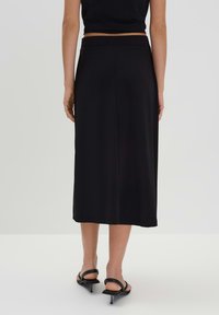 Jupe midi noire en tissu lisse, dotée d'une taille ajustée et d'une silhouette droite et fluide. Portée avec des sandales à talons noires minimalistes.