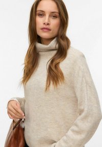 Bež pulover z visoko ovratnico s mehko teksturo, dolgimi rokavi in rebrastim vzorcem, združen z rjavo torbico čez ramo.