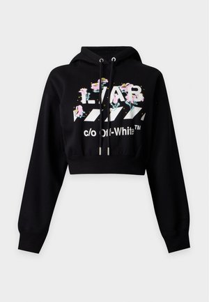 Jeune femme portant un sweat à capuche court noir et un pantalon noir à cordon avec motif floral et texte, debout sur fond blanc.