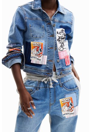 Chaqueta vaquera y vaqueros con parches de Mickey Mouse, llevados por una persona con una mano en el bolsillo y dos anillos de plata en la otra mano.