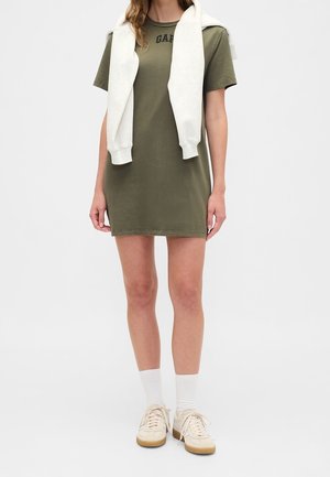 Personne portant une robe T-shirt vert olive, des chaussettes blanches, des baskets beiges et un sweat blanc drapé sur les épaules.