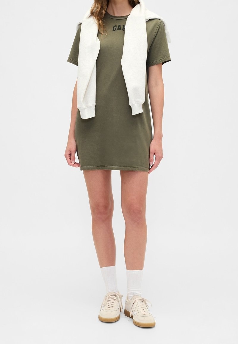 Personne portant une robe T-shirt vert olive, des chaussettes blanches, des baskets beiges et un sweat blanc drapé sur les épaules.