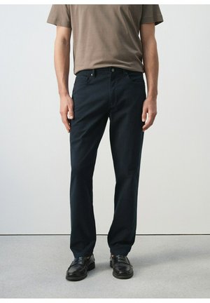 Hombre vistiendo pantalones rectos negros, mocasines de cuero negros y una camisa beige de manga corta, de pie contra un fondo liso.