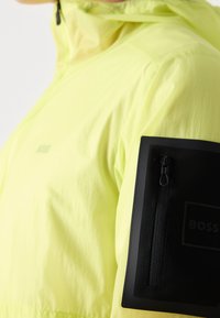 Veste jaune vif à col montant, dotée d'une poche zippée noire sur le haut du bras avec un logo de marque visible.
