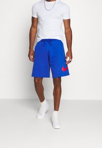 Blå sportshorts med snörmidja, med en röd Nike-logotyp på höger ben. Bärs med en vit t-shirt och vita sneakers.