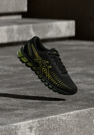 Czarny i zielony but sportowy z żółtymi akcentami unoszący się nad betonowymi schodami, podkreślający amortyzowaną podeszwę i smukły design.