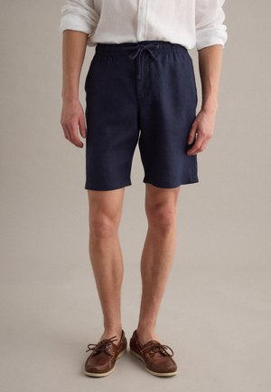 Mann trägt marineblaue Shorts mit Kordelzug, braune Bootsschuhe und ein weißes Langarmhemd mit hochgekrempelten Ärmeln vor einem einfarbigen Hintergrund.