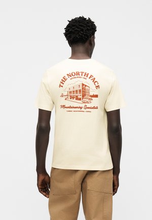 Mand iført beige North Face t-shirt med orange grafik og teksten "Mountaineering Specialists" på ryggen, kombineret med lysebrune bukser.