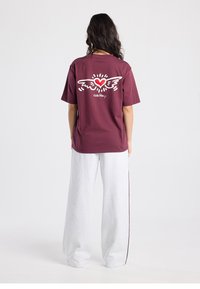 T-shirt en coton bordeaux avec un motif de cœur ailé blanc dans le dos, associé à un pantalon de survêtement ample gris clair avec des bandes latérales foncées.