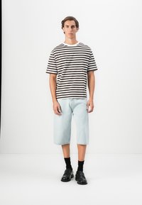 Stripet bomull t-skjorte i brun og hvit, kombinert med lyseblå denimshorts. Modellen har på seg sorte loafers og sorte ankelstrømper.