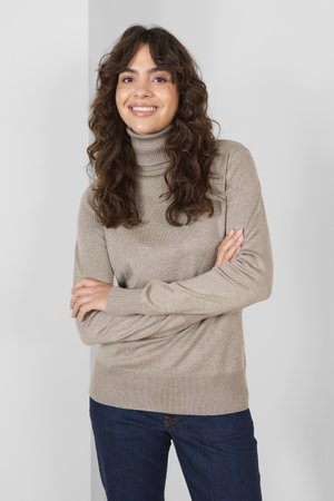 Beige coltrui met geribde manchetten en zoom, gemaakt van zacht materiaal. Het model draagt het met donkerblauwe jeans en staat tegen een lichte achtergrond.