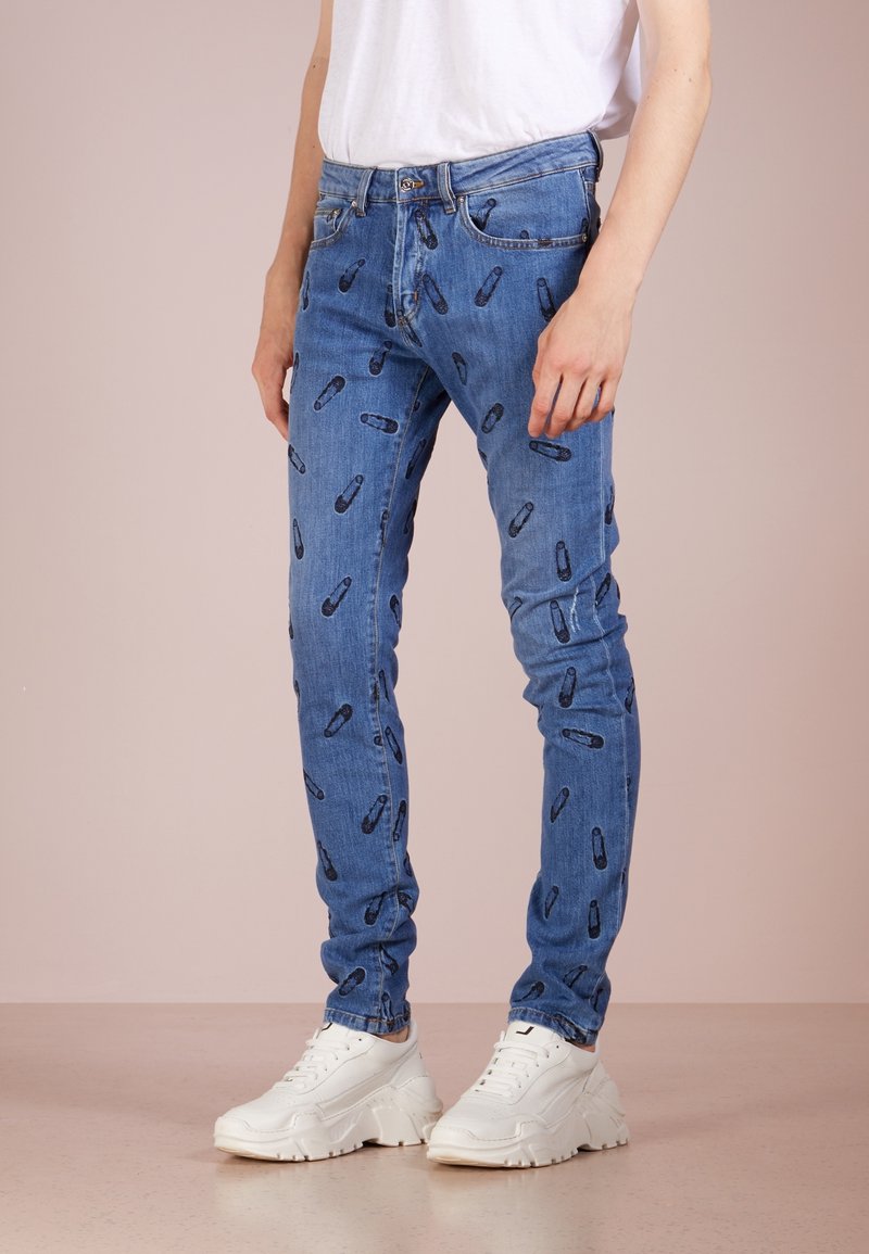Modré džínové skinny jeans s opakujícím se vzorem bezpečnostních špendlíků, pěti kapsami a zapínáním na knoflík, doplněné o bílé tenisky.