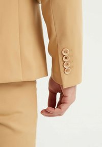 Manche de veste beige comportant quatre boutons crème avec des détails de couture. Texture lisse avec un léger éclat, coupe ajustée visible.