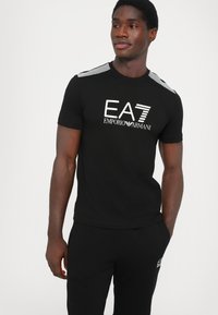 Juodas trumpomis rankovėmis marškinėliai su baltu "EA7 Emporio Armani" logotipu, turintys dryžuotas akcentus ant pečių. Minkšta medvilnės medžiaga.