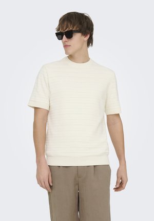 Jeune homme portant des lunettes de soleil noires, un haut tricoté crème à manches courtes, et un pantalon beige, posant devant un fond blanc uni.