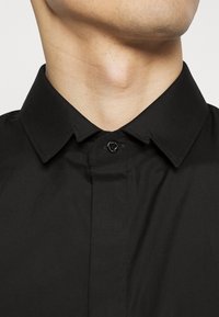 Camicia nera con bottoni, dotata di un colletto strutturato, con una chiusura a un bottone sullo scollo e una texture di tessuto liscia.