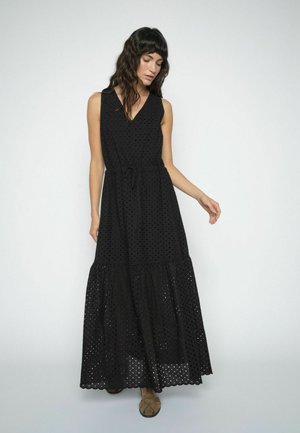 Žena oblečená v čiernych maxi šatách bez rukávov s očkovaným vzorom a viazaním v páse, stojaca pred jednoduchým svetlým pozadím.