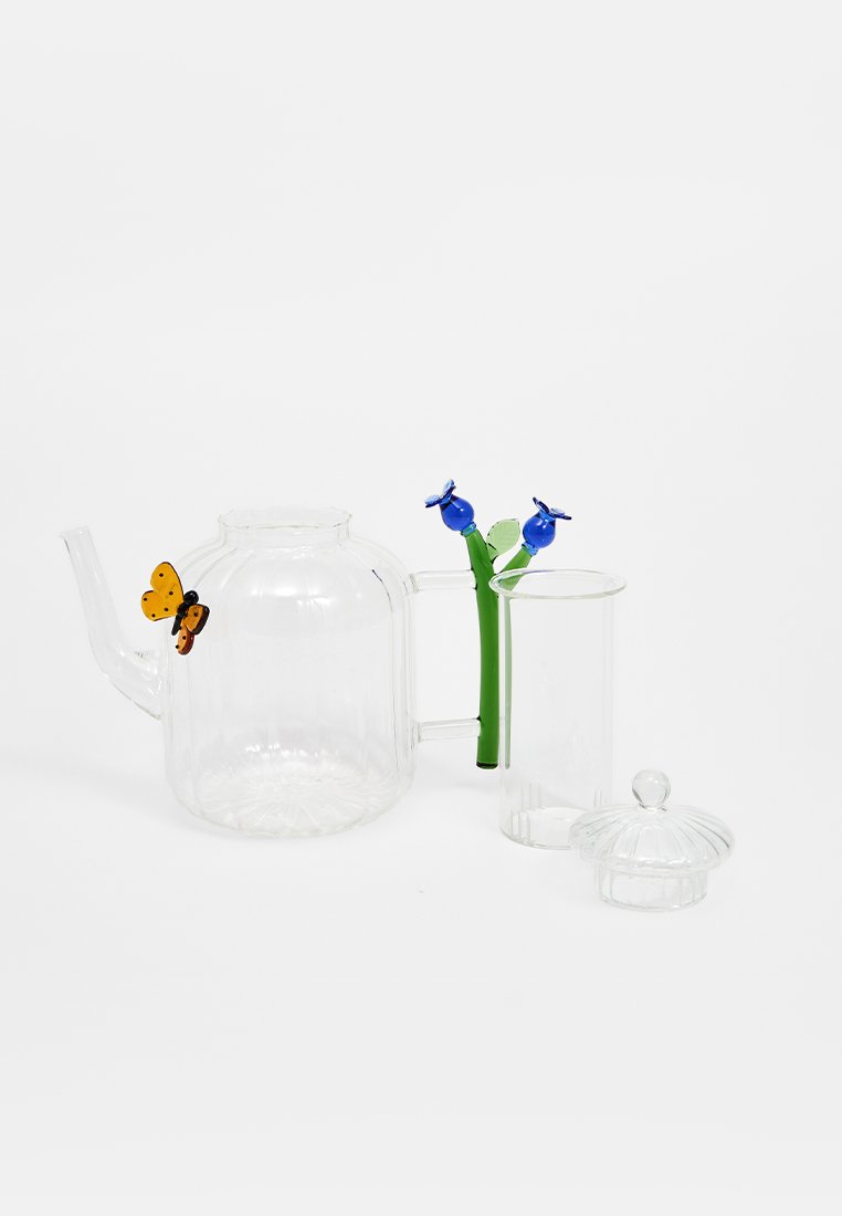 Przezroczysty szklany czajnik z szerokim dnem i smukłym dzióbkiem, z zielonym uchwytem ozdobionym niebieskimi kwiatami, w zestawie z przezroczystą filiżanką i pokrywką.