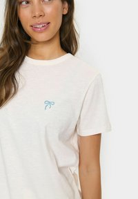 Camiseta de manga corta en color crema claro, hecha de una tela suave, con un pequeño lazo azul bordado en el pecho. Cuello redondo y corte relajado.