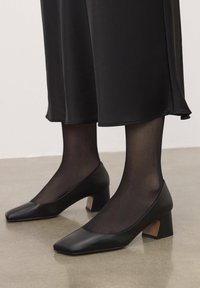 Scarpe in pelle nera con tacco medio, punta quadrata e dettaglio in cuoio marrone texturizzato sul tacco, indossate con collant neri trasparenti e una lunga gonna nera.