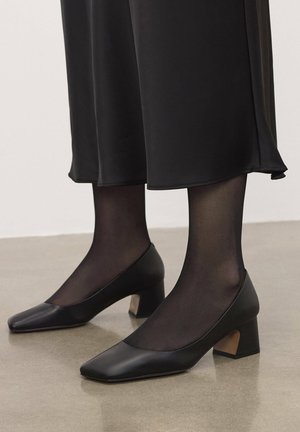 Scarpe in pelle nera con tacco medio, punta quadrata e dettaglio in cuoio marrone texturizzato sul tacco, indossate con collant neri trasparenti e una lunga gonna nera.