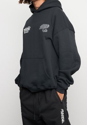 Persona che indossa una felpa nera con cappuccio con il testo bianco "Wrongly Accused 2024" e pantaloni neri con il logo bianco "Multiply Apparel", mani nelle tasche.