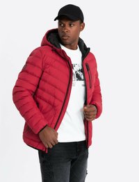 Doudoune rouge avec capuche, présentant un matelassage horizontal, des accents noirs et une poche zippée. Portée sur une chemise blanche et un jean noir.