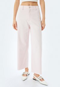 Jean large rose clair à taille haute, à cinq poches et ourlet brut. Associé à des chaussures à talons ouverts blanches à bout pointu.