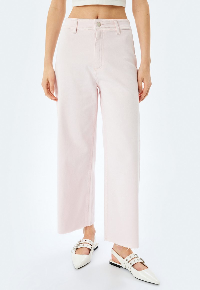 Jean large rose clair à taille haute, à cinq poches et ourlet brut. Associé à des chaussures à talons ouverts blanches à bout pointu.