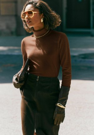 Femme portant un col roulé marron, une jupe noire, des gants en cuir, de grandes lunettes de soleil jaunes, tenant une pochette noire, marchant en extérieur sous le soleil.