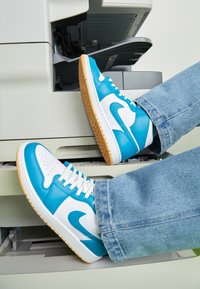 Blue and white sneakers with a perforated toe, rubber sole, and logo accents, resting on a copier in a light-colored setting.