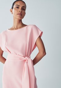 Vaaleanpunainen mekko rentoa istuvuutta, venepääntie ja lyhyet hihat. Sisältää solmittavan vyötäröyksityiskohdan ja pehmeän, valuvan kankaan tekstuurin.