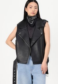 Črn brezrokavnik iz umetnega usnja z asimetričnim zadrgo, širokimi klamfami in obliko z beljenjem, v kombinaciji z črn vtičnikom in topom s pikami.