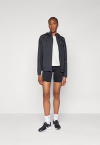Veste légère noire avec capuche et fermeture éclair à l'avant, associée à un haut blanc et un short noir, portée avec des baskets noires et des chaussettes blanches.