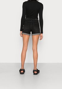 Svarta jeansshorts med uppvikta fållar, figurnära passform, bakfickor och kombinerade med svarta sandaler, visade mot en neutral bakgrund.