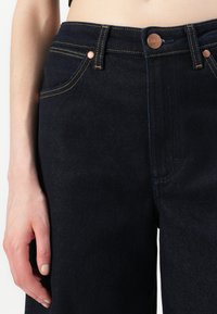 Close-up of a person wearing dark blue denim jeans with yellow stitching, copper rivets, and a button closure, hand resting by side.