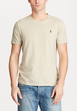 T-shirt beige à manches courtes en coton avec un col rond ; il présente un petit logo brodé sur la poitrine gauche.