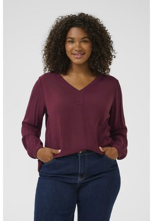 Bluza lungă burgundy cu mânecă lungă și decolteu în V, având o textură subtilă, un croi lejer și manșete pliate. Asortată cu blugi bleu închis.