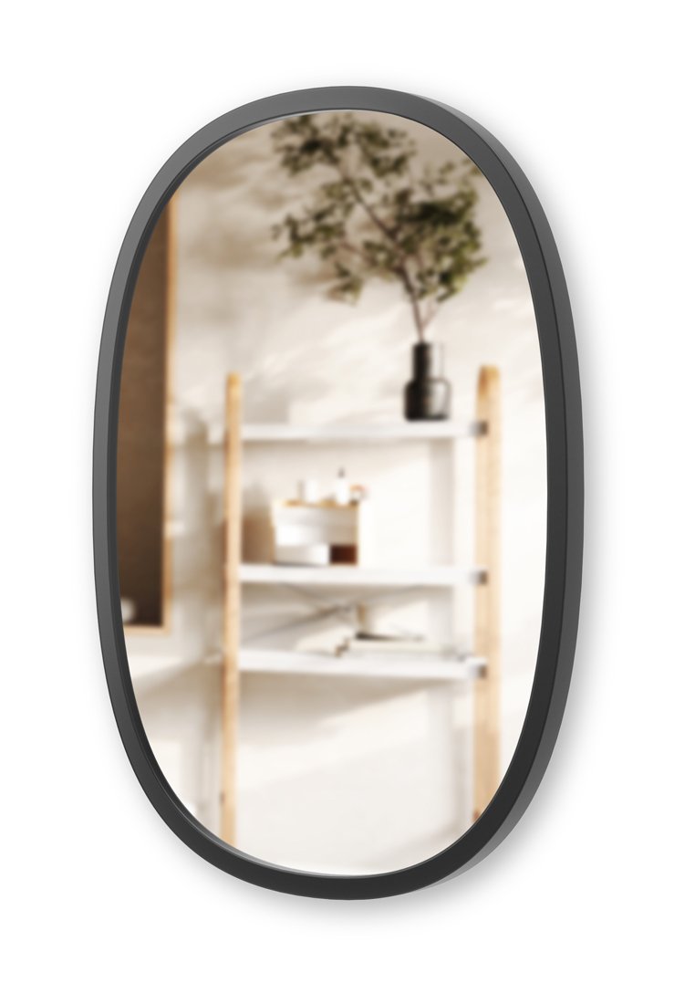 Specchio ovale con cornice nera che riflette una scaffale a scala in legno con ripiani bianchi che ospitano piccoli oggetti decorativi e un rametto verde in un vaso nero.