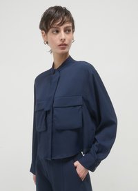 Navy blue cropped jacket with a high collar, large front pockets, and long sleeves. The fabric is smooth, featuring a subtle sheen.