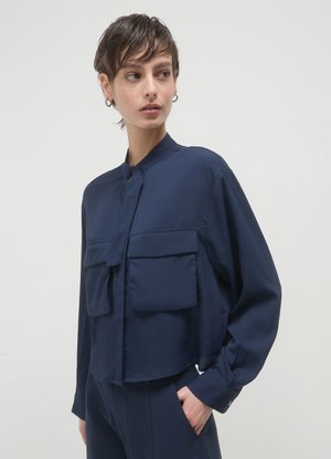 Marineblaues, kurzes Jackett mit hohem Kragen, großen vorderen Taschen und langen Ärmeln. Der Stoff ist glatt und hat einen subtilen Glanz.