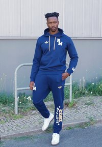 Hoodie azul marinho com detalhes dourados e logótipo branco, combinado com calças de treino correspondentes que apresentam detalhes impressos, e ténis brancos.