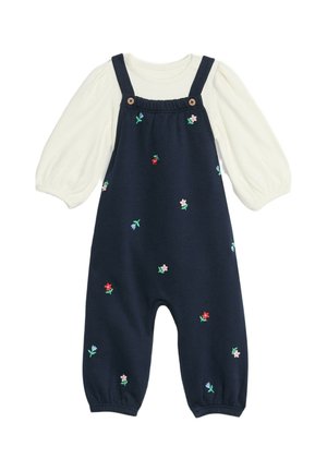Salopette en coton marine avec des fleurs brodées en rose, rouge et vert. Dotée de bretelles ajustables, de boutons en bois et d'un haut à manches longues crème en dessous.