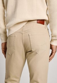 Jeans de algodón beige con dos bolsillos traseros y un parche de cuero. Presentan un corte Slim y un diseño de costura clásico y sutil.